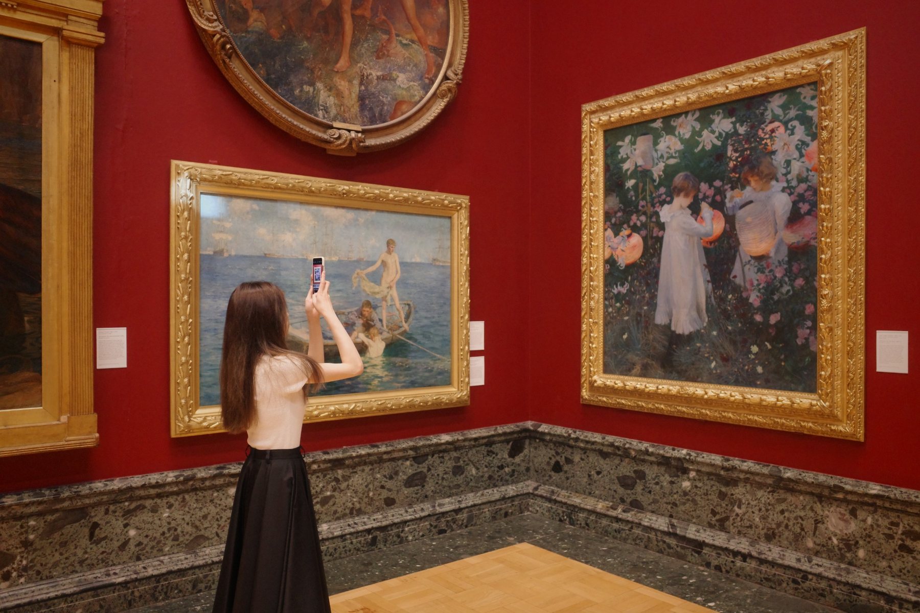 A person photographing paintings in a red-walled gallery.